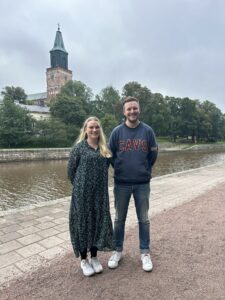 Kuvassa Matkalla-hankkeen valmentajat Linda ja Perttu Turun jokirannassa. Heidän taustallaan näkyy joki ja Turun tuomiokirkko.